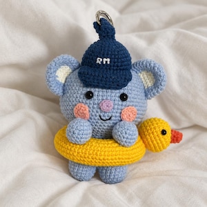 Op de afbeelding: Een lichtblauwe gehaakte koala-sleutelhanger met een donkerblauwe pet waarop de witte letters RM aan de voorzijde zijn geborduurd. De koala heeft roze wangen, een paarse neus en zit in een felgele, eendvormige zwemband.