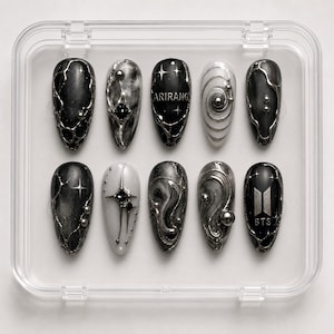 Pode incluir: Um conjunto de dez unhas postiças em formato amendoado num estojo de plástico transparente. As unhas apresentam designs metálicos em preto, branco e prata, incluindo esferas prateadas em relevo, estrelas, padrões espirais e o texto ARIRANG e BTS.