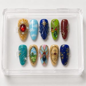 Puede incluir: Un conjunto de diez uñas postizas en forma de almendra presentadas en una caja de plástico transparente. Cada uña presenta diseños únicos que incluyen paisajes pintados a mano, marcos de filigrana dorada, pedrería, perlas y motivos celestiales.