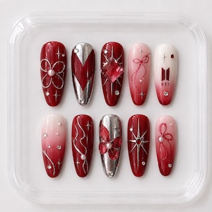 May include: A set of ten press-on nails featuring a red, white, and silver color palette. Designs include metallic silver accents, red rhinestones, small pearls, star-shaped embellishments, a red bow illustration, and the BTS logo in black.