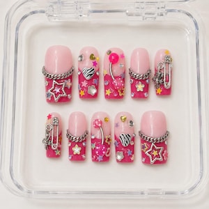 Könnte beinhalten: Ein Set aus zehn künstlichen Fingernägeln in Pink und Hellrosa mit French-Tip-Design. Die Nägel sind mit silberfarbenen Metallketten, kleinen Sicherheitsnadeln, sternförmigen Anhängern, Strasssteinen und herzförmigen Akzenten verziert.