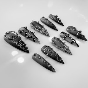 May include: A selection of grey nail covers with metal detailing, including rings, studs, and decorative elements. The nail covers are arranged on a white surface. The designs have a gothic, punk aesthetic.