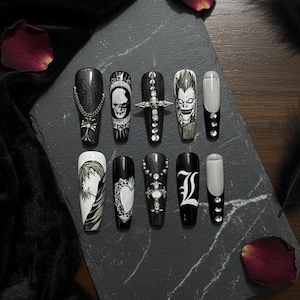 May include: A set of ten long, coffin-shaped artificial nails. Each nail is black, with various designs including a skull, cross, and anime-style character. Some nails feature silver chain and gem accents.