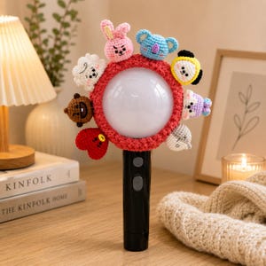 May include: A black light stick featuring a circular white globe encased in a textured red crochet cover. The cover is adorned with eight small, colourful, crocheted character figures arranged around the perimeter, including a pink rabbit and blue koala.