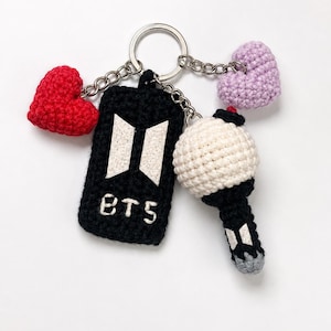 Puede incluir: Un llavero de metal con tres dijes de ganchillo: un corazón rojo, un corazón morado claro y una etiqueta rectangular negra con el logotipo y el texto BTS en blanco. Un cuarto dije es una esfera blanca de ganchillo sobre un mango negro.