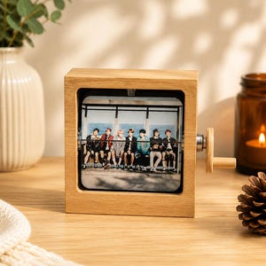 May include: A square light-colored wooden frame containing a rotating display panel that features a printed photograph of seven individuals sitting together on a bench. A small wooden crank handle is attached to the right side of the frame.