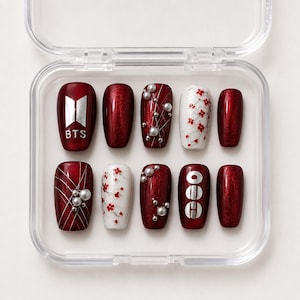 Pode incluir: Um conjunto de dez unhas postiças num estojo de plástico transparente. As unhas apresentam um acabamento vermelho metálico profundo, detalhes em branco com padrões florais vermelhos, linhas prateadas e pequenas pérolas brancas.