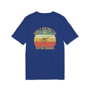 May include: A royal blue short-sleeve t-shirt featuring a graphic of a small airplane centered within a distressed sunset circle. The text reads EASILY DISTRACTED BY PISTON ENGINES in a yellow sans-serif font arched above and below the airplane graphic.
