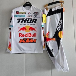 Könnte beinhalten: Weißes Motocross-Trikot- und Hosen-Set mit schwarzen und orangefarbenen Akzenten. Das Trikot zeigt Logos von Thor, Red Bull und KTM. Die Hose hat ein schwarz-weißes Design mit dem Thor-Logo. Der Text auf dem Bild lautet "FREE NAME & NUMBER" und "CUSTOMIZABLE".