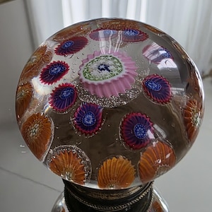 May include: A clear glass paperweight with embedded colorful floral designs. The paperweight features orange, purple, and pink flowers, with a decorative base. The glass sphere reflects the surrounding environment.