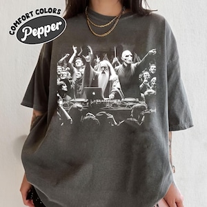 May include: Dark grey t-shirt with a graphic of a crowd at a concert. The graphic features a wizard and another figure at a DJ setup, with a laptop. The text "COMFORT COLORS Pepper" is in a white oval on the upper left.