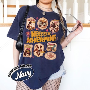 May include: Navy blue t-shirt with a graphic design featuring cartoon characters and the text "NEEEEEW ACHIEVEMENT!". The design includes various badges with phrases like "THIS OUT IS AN RAGE" and "MONGO IS AWESOME".
