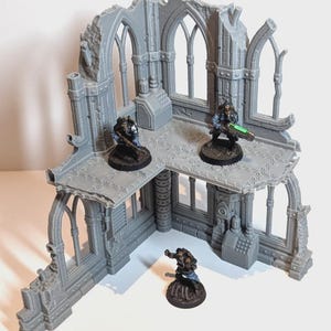 May include: A gray, multi-level miniature building with arched windows and battle damage. Three miniature figures are positioned on the structure. The building has a textured surface and appears to be made of plastic. The figures are painted with detail.