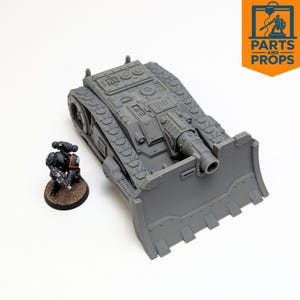 May include: A grey, 3D-printed miniature tank model with a large blade at the front and detailed track treads. A small, painted figurine of a soldier is positioned next to the tank. The image includes the logo for "Parts and Props".