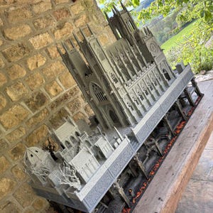 May include: A detailed gray miniature cathedral model with intricate architectural details, including spires, arched windows, and decorative carvings. The model sits on a wooden surface, with a brick wall in the background.
