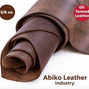 Puede incluir: Cuero curtido al aceite, de color marrón oscuro, enrollado. La imagen muestra la textura y las capas del cuero, con una etiqueta de 5/6 oz. El texto "Abiko Leather industry" se encuentra en la parte inferior.