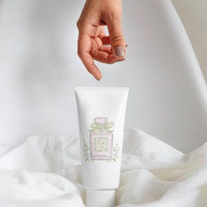 Puede incluir: Un tubo blanco con una ilustraci&oacute;n de acuarela de un frasco de perfume en rosa y verde, atado con un lazo verde. Una mano est&aacute; colocada encima del tubo. El texto en la parte superior dice "Watercolor Collection" y en la parte inferior "Collection by Pati Paints".