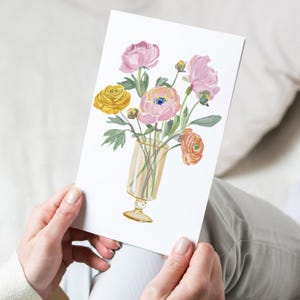 May include: A greeting card with a watercolour illustration of a floral bouquet in a clear glass vase. The flowers are pink, yellow, and orange, with green leaves. The card is held by a person.