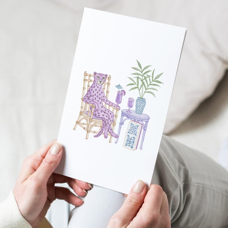 K&ouml;nnte beinhalten: Eine wei&szlig;e Gru&szlig;karte mit einer farbenfrohen Illustration eines lila gefleckten Leoparden, der auf einem beigen Bambusstuhl sitzt. Der Leopard h&auml;lt einen lila Cocktail mit einem blauen Schirmchen. Daneben steht ein kleiner Tisch mit einer Pflanze.