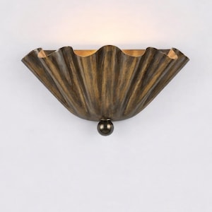 May include: A wall sconce with a scalloped, fan-shaped design. The fixture has a bronze finish with a dark, textured appearance. A small, dark sphere hangs below the light. The interior of the sconce has a golden hue.