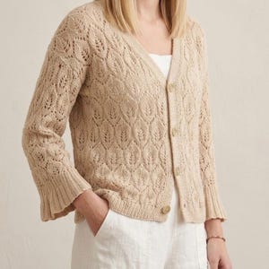 May include: A beige cardigan with a V-neck and button closure. The knitwear features a leaf pattern and flared sleeves. The person is wearing white pants and has their hand in their pocket. The cardigan is a neutral color.