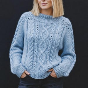 May include: A light blue long-sleeve knit sweater featuring a textured cable knit pattern on the front and sleeves, a ribbed crew neckline, and ribbed cuffs. The garment has a soft, fuzzy appearance with intricate vertical braided cable details.
