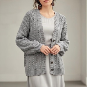 May include: A gray cable-knit cardigan with a button-up front. The sweater has a relaxed fit with long sleeves and a textured pattern. The buttons are a dark brown color. The cardigan is worn over a light gray dress.