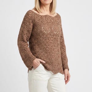 May include: A brown crochet sweater with a scalloped neckline and long sleeves. The sweater features a floral pattern throughout and is paired with white pants. The crochet design is visible, showcasing the intricate details of the garment.