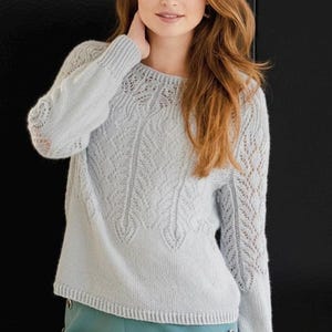 May include: A pale blue knitted jumper with a round neck and long sleeves. The jumper has a detailed leaf pattern across the front and sleeves. The sleeves feature an openwork design. The garment is made of a soft, light-coloured yarn.