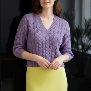 May include: A lilac V-neck sweater with a cable knit pattern. The sweater features long sleeves and ribbed detailing at the hem and cuffs. The model is wearing a yellow skirt. The sweater is made of a soft, comfortable material.