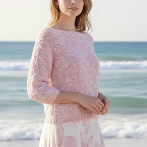 Può includere: Un maglione rosa chiaro con scollo a barchetta e maniche a tre quarti. Il maglione presenta un motivo a maglia traforata strutturata. La modella indossa una gonna rosa chiaro e bianca. Lo sfondo è una spiaggia con l'oceano.