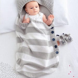 Puede incluir: Un bebé en un saco de dormir de punto a rayas grises y blancas con una capucha con diseño de cara de gato. El saco tiene cierres de botón y un cordón azul oscuro. Un par de patucos a juego están junto al saco.