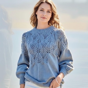 Może przedstawiać: Jasnoniebieski sweter z detalicznym wzorem liści na piersi i ramionach. Sweter ma okrągły dekolt i długie rękawy z lekkim pufem. Modelka ma na sobie srebrną bransoletkę i białe spodnie.