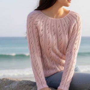 May include: A pale pink knitted sweater with a round neck and long sleeves. The sweater has a detailed openwork design and is paired with blue jeans. The background shows a beach and the sea.