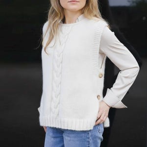 May include: Cream-colored cable knit sweater vest with a button-up side detail. The vest features a central cable knit design and is paired with a long-sleeved, light-colored shirt and blue jeans. The overall style is casual and comfortable.