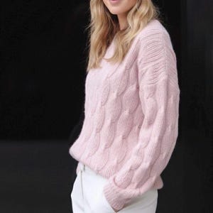 May include: A light pink cable-knit sweater with a textured pattern. The sweater has long sleeves and a round neckline. The person is wearing white pants. The background is black.