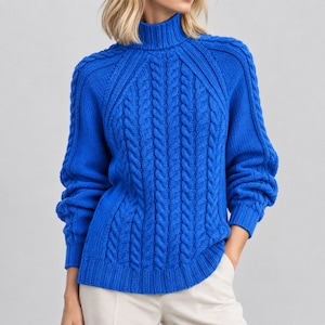 Puede incluir: Un suéter de punto trenzado en color azul real vibrante con cuello alto, mangas largas con puños acanalados y un prominente patrón de trenzas verticales en la parte delantera y los hombros, combinado con pantalones claros.