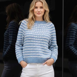 May include: A light blue and white striped sweater with a crew neck. The model is wearing white trousers. The background is a dark, reflective surface. The sweater has long sleeves.