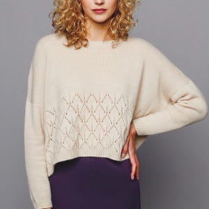 May include: A cream-coloured sweater featuring a diamond pattern across the midsection. The sweater has a crew neck and long sleeves. The bottom hem is cropped. The sweater is paired with a purple skirt.