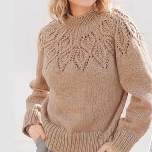 Puede incluir: Un suéter tejido de color beige con cuello y puños acanalados. El canesú presenta un diseño decorativo de hojas caladas. El suéter tiene mangas largas y un dobladillo acanalado. El estilo es cómodo y cálido.