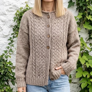 May include: A beige cable-knit cardigan with a button-up front. The sweater has a textured pattern and a round neckline. The long-sleeved cardigan is made of a soft, warm material, suitable for cooler weather. The buttons are a light brown colour.