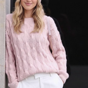 May include: A light pink cable knit sweater with a crew neck. The sweater is paired with white pants. A light pink purse with a gold chain strap is visible. The person is wearing a light pink beanie.