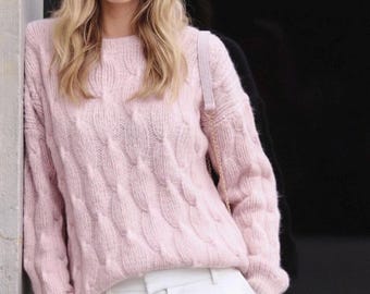 Könnte beinhalten: Ein hellrosa Zopfmuster-Pullover mit Rundhalsausschnitt. Der Pullover hat ein strukturiertes Muster und wird mit einer weißen Hose kombiniert. Eine hellrosa Handtasche mit goldenem Kettenriemen ist sichtbar.