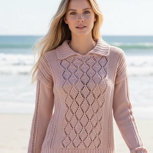 May include: A light pink collared sweater with a diamond-patterned front. The long-sleeved sweater has ribbed cuffs and a ribbed hem. The model is standing on a beach with the ocean in the background.