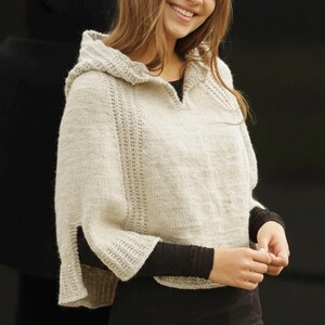 May include: A light beige hooded poncho with a split sleeve design. The poncho is made of a knitted material and features a textured pattern along the sleeves and hood. The model is wearing a black long-sleeved top underneath.