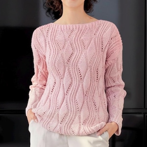 May include: A pale pink, long-sleeved jumper with a boat neck. The jumper has a detailed, open-weave pattern with vertical, wave-like designs. The sleeves and body have a ribbed texture. The model is wearing light-coloured trousers.
