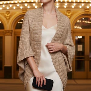 Può includere: Uno scialle beige a maglia con maniche ampie è indossato sopra un vestito bianco. Lo scialle ha un aspetto strutturato. Una piccola pochette nera è tenuta in mano. Lo sfondo presenta un edificio con dettagli dorati e luci.