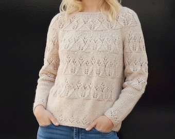 May include: A beige, long-sleeved sweater with intricate leaf-like patterns. The sweater has a round neckline and is made of a soft, textured yarn. The sleeves are long and fitted, and the overall design is elegant and detailed. The person is wearing blue jeans.
