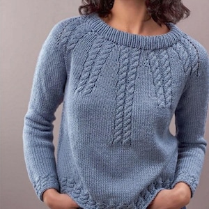 Può includere: Un maglione lavorato a maglia blu chiaro con scollo tondo e maniche lunghe. Il maglione presenta un motivo a trecce sul davanti e lungo le spalle, con polsini e orlo a coste. La persona indossa il maglione con le mani in tasca.