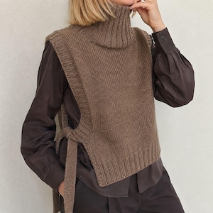 Può includere: Un gilet in maglia marrone con collo alto a lupetto, bordi a coste su giromanica e orlo, e lacci laterali che chiudono i lati aperti. Il capo è indossato sopra una camicia marrone scuro a maniche lunghe con polsini abbottonati.